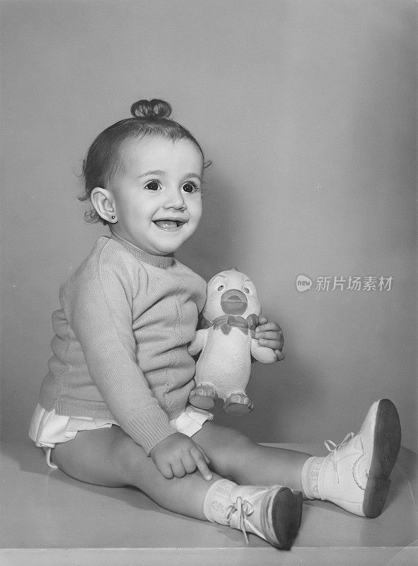 黑白照片拍摄于60年代，小女孩摆姿势拿着手机看向别处，手里拿着玩具