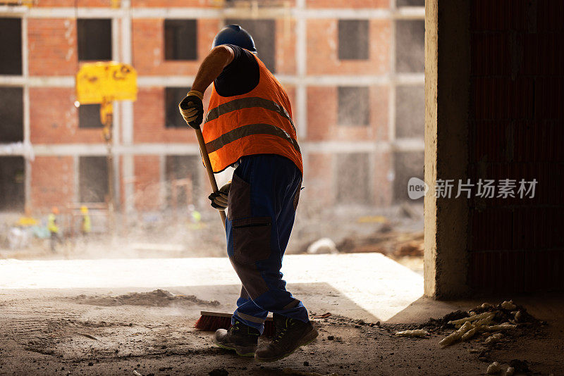 身穿橙色反光背心的建筑工人，正在用扫帚清理工地上的灰尘