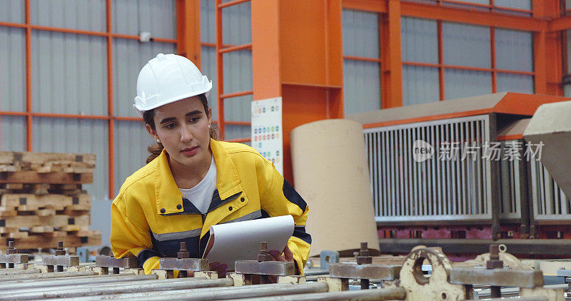 年轻的拉丁技术员妇女戴着安全安全帽，拿着剪贴板，在工业工厂边走边检查库存