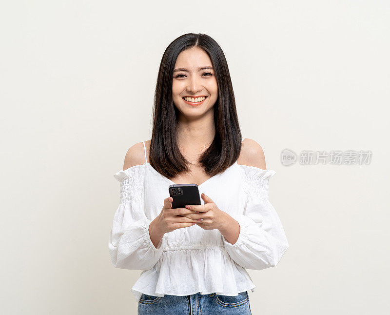 年轻漂亮的亚洲女子站在孤立的白色背景上使用智能手机。兴奋年轻漂亮的亚洲人用手机网购支付。用智能手机玩游戏