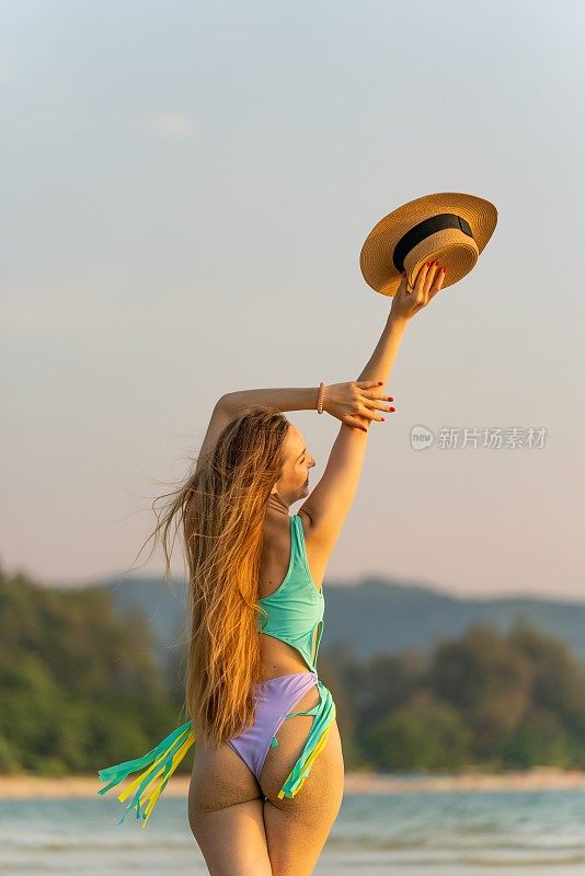 一个美丽的白人女人在比基尼性感的背影在海滩上拿着一顶沙滩帽。女士享受海风和日落美景