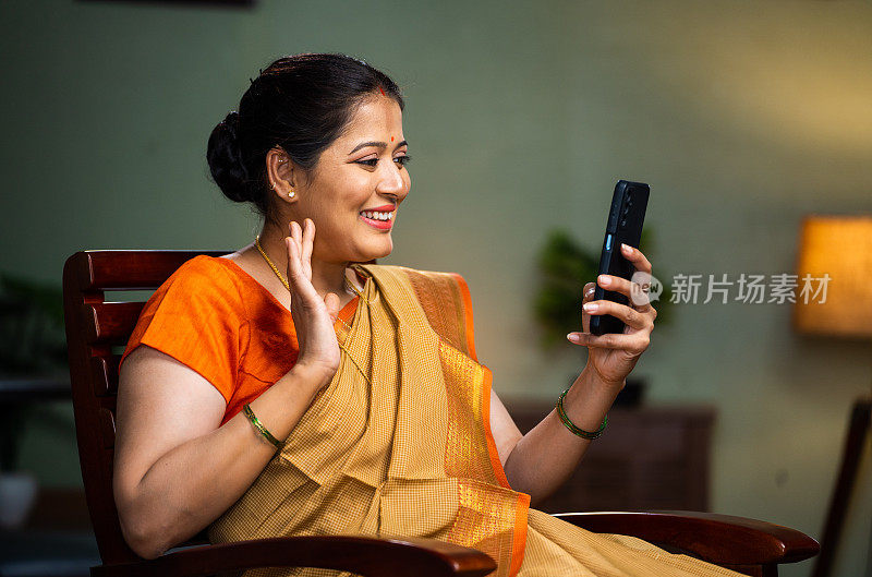 快乐的印度中年妇女坐在家里的椅子上忙着用手机打视频电话——技术、放松和联系的概念