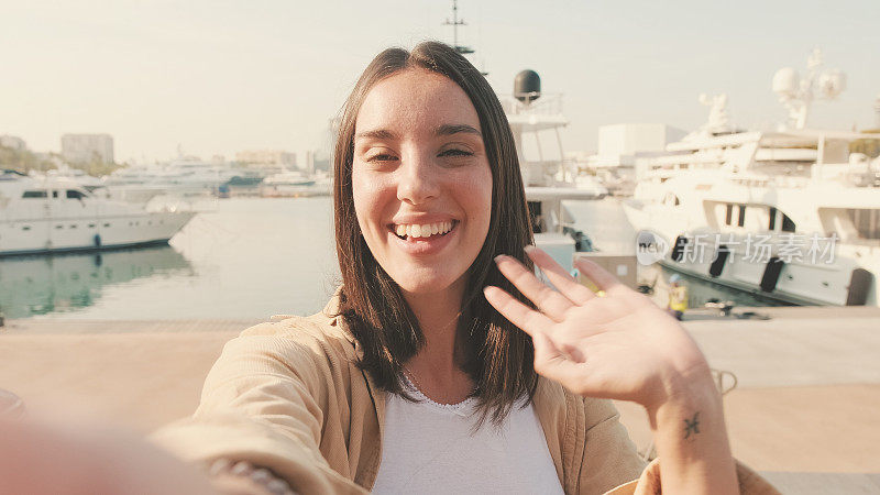 近距离，在欧洲城市的游艇背景下，站在港口用手机打视频电话的旅行者女孩挥舞着手掌打招呼