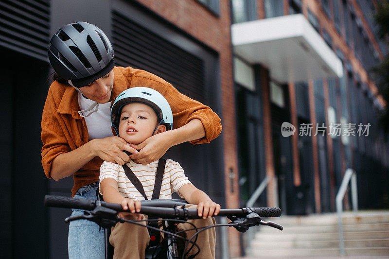 母亲把儿子的自行车头盔系在头上，把他抱在儿童自行车的车架上，放在座位上。母亲带着年幼的孩子骑着自行车在城市里穿梭。