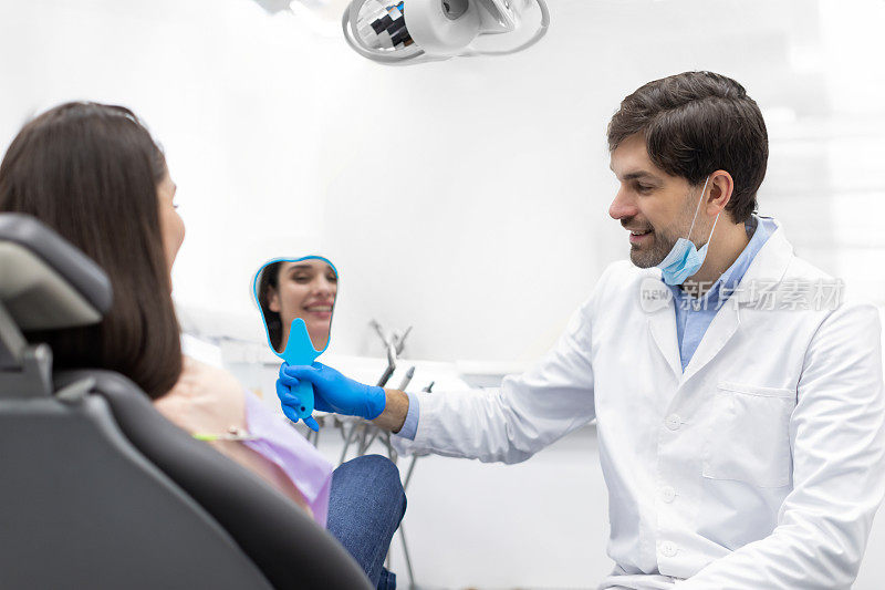 妇女在诊所接受牙科治疗后对着镜子检查自己的牙齿，男牙医向女病人展示治疗结果