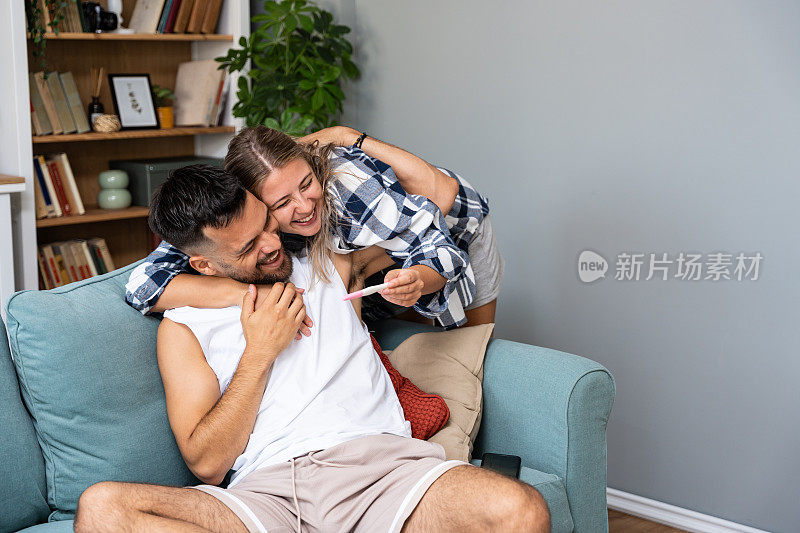 年轻幸福的夫妇，怀孕的女人在他们的公寓里向男人展示怀孕测试的阳性结果。新婚夫妇很棒的新闻概念。