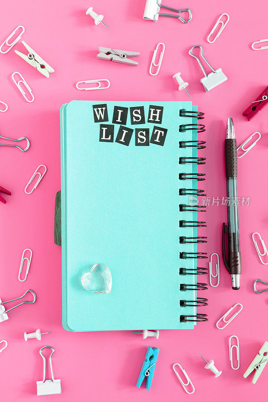 一个愿望清单。记事本弹簧与蓝色页在粉红色的背景。办公用品杂乱摆放。夹子和文书按钮混淆。上面的照片很漂亮。