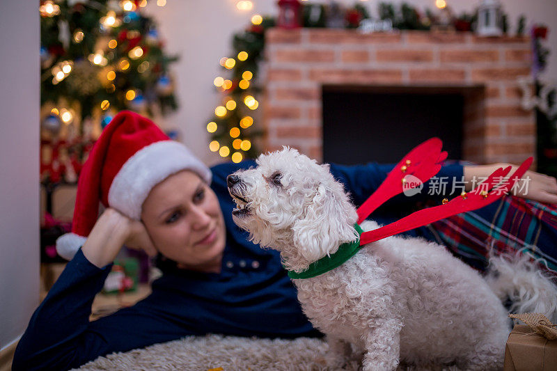 有吸引力的时髦的年轻女人圣诞节与她的狗坐在装饰的壁炉和树轻拍它的头与幸福的微笑。