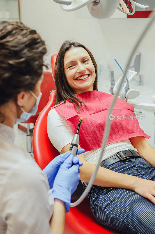 一个年轻女子坐在牙医的椅子上预约医生。这段时间她很担心害怕，然后她看着镜头。医生-一个穿着医疗服的好女人，展示了一个牙科灯并解释了她为什么是。