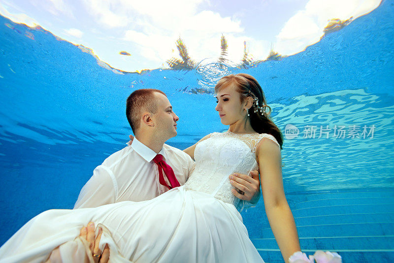 在一个阳光明媚的日子里，新郎和新娘在游泳池的水下。新郎把新娘抱在怀里，年轻人互相看了看。肖像。的概念。在水下婚礼。横向视图