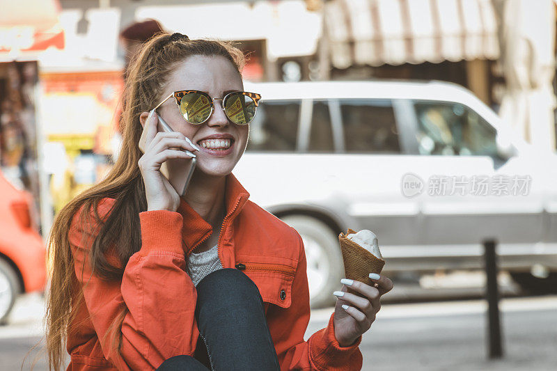 吃着冰淇淋的迷人女人正坐在城市街道上用智能手机聊天