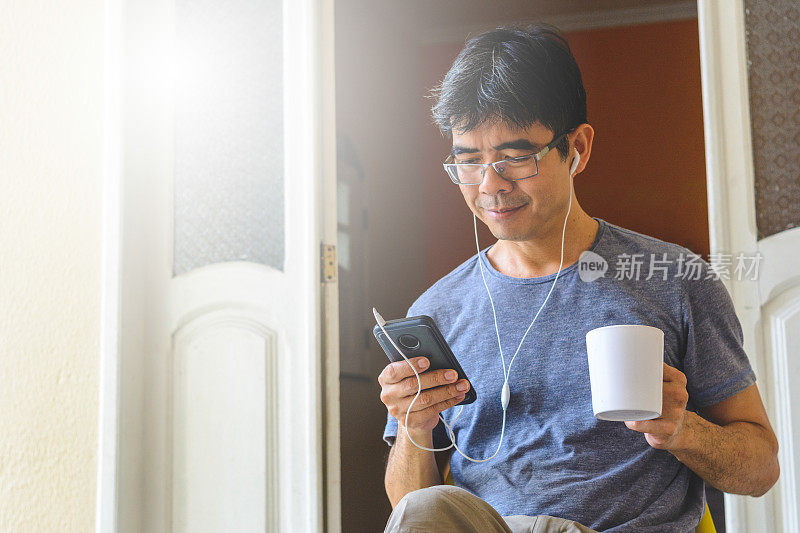 成熟的亚洲男人使用智能手机和耳机，并拿着一杯咖啡或茶