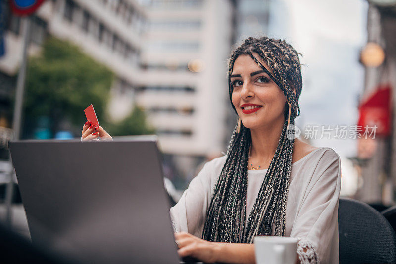 一位女士坐在路边的咖啡馆里，拿着信用卡使用笔记本电脑