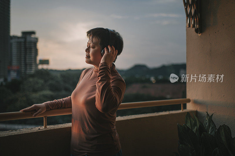 日落时分，一名亚裔华人中年妇女站在阳台上，戴着无线耳机听音乐