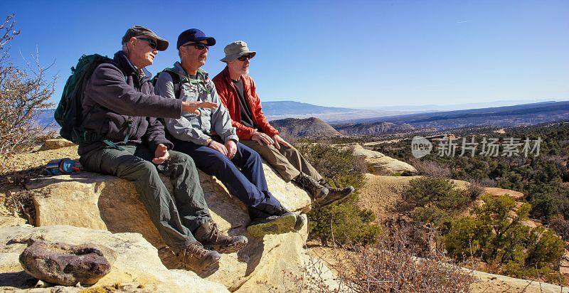 三个中年男子坐在科罗拉多西部沙漠山上的一块岩石上，一边聊天一边欣赏风景