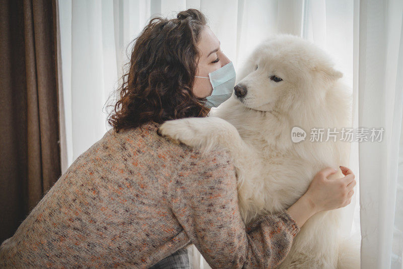 年轻女子戴着医用口罩，带着狗站在窗边