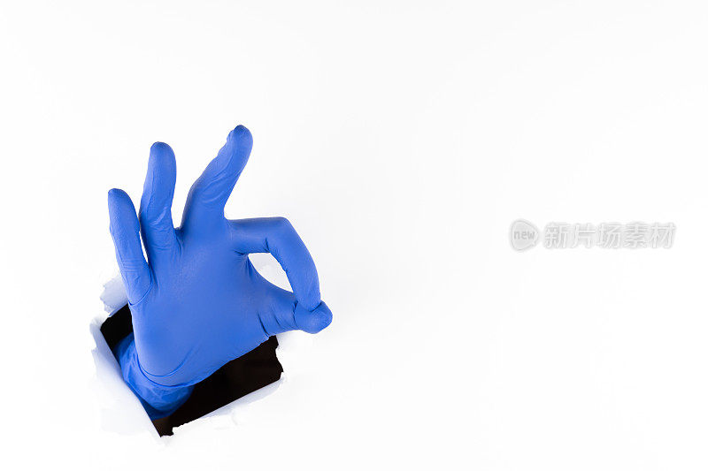 一名妇女戴着蓝色橡胶手套的手从白纸上的破洞中伸出手指，表示没事。一切都会好的。