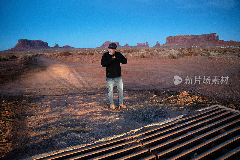 日落时分，在亚利桑那州北部纳瓦霍地区纪念碑谷部落公园雄伟的山头前，一名成熟的西班牙男子穿着外套，戴着手套取暖