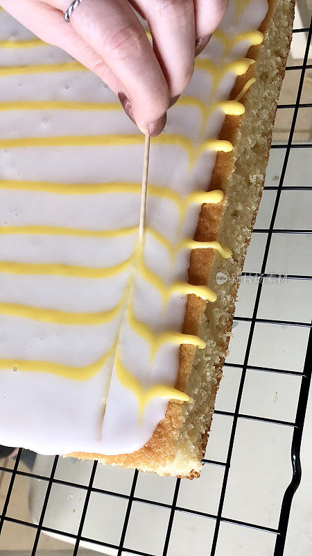 不认识的人的手用鸡尾酒棒画一个黄色的羽毛图案，柠檬味的糖霜通过白色方糖糖霜，自制的柠檬托盘烤蛋糕在金属冷却架，高架视图