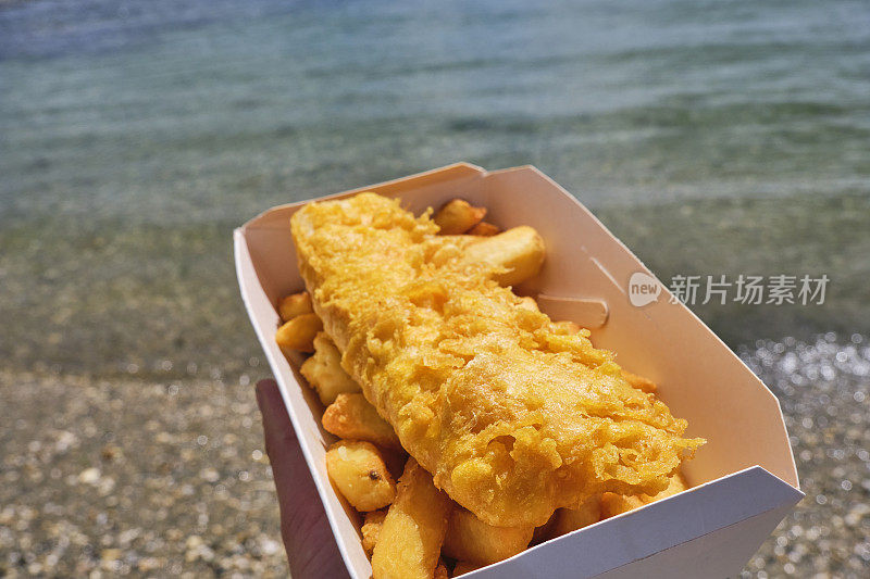 在康沃尔圣莫斯清澈浅海的海岸线上，一只手拿着一盒炸鱼薯条。