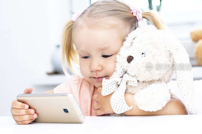 女孩拿着玩具兔用手机，用智能手机打视频电话，和亲戚聊天，女孩坐在家里，上网电脑摄像头，打视频电话。