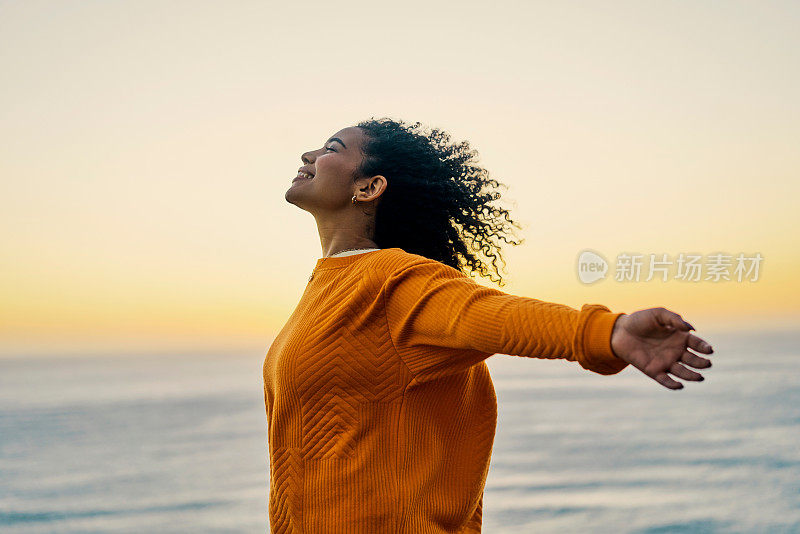 自由，自由和快乐的黑人女性在日落时分在海滩庆祝自由，积极而兴奋。年轻女性享受海滩度假和精神力量，同时站着呼吸新鲜空气