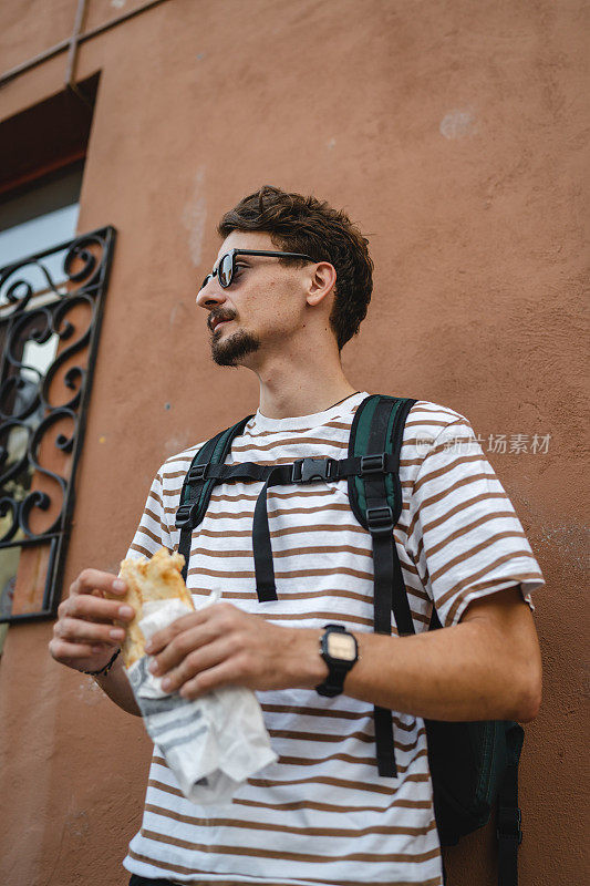 一个男人年轻的成年现代白人男性在城市里在阳光灿烂的日子里站着吃三明治快餐概念城市生活复制空间游客在建筑墙前吃真人画像
