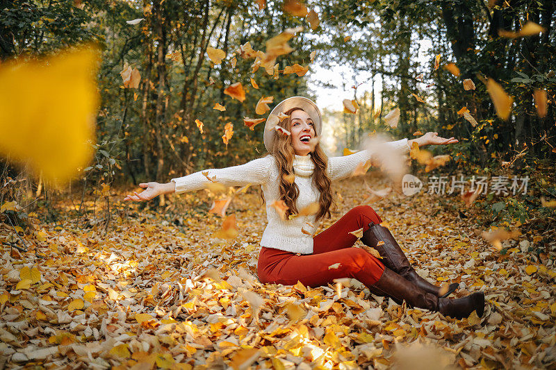 在阳光灿烂的日子里，微笑着的卷发女人在秋天的森林里扔树叶