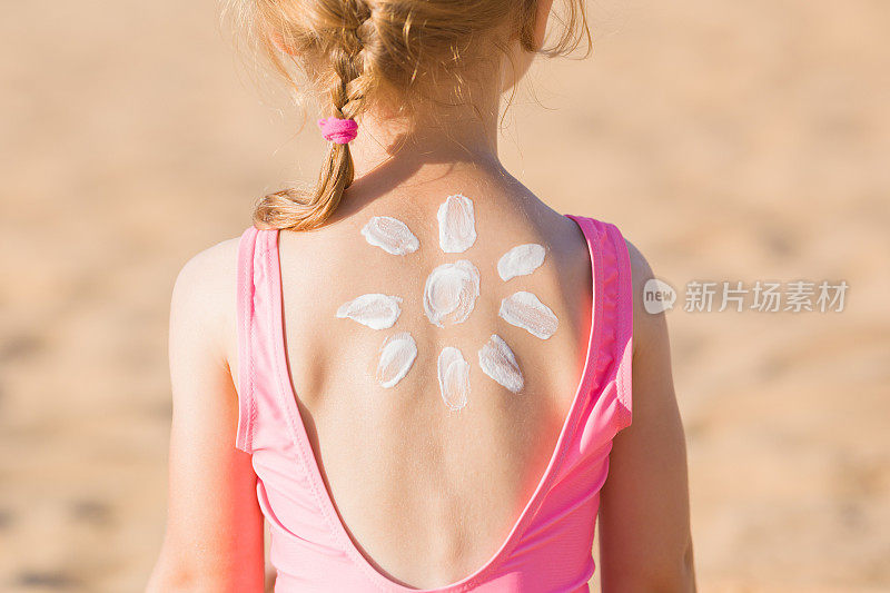 太阳形状创建从白色防晒霜小女孩背部的沙子背景。皮肤的保护。在烈日炎炎的沙滩上进行安全日光浴。后面的观点。特写镜头。