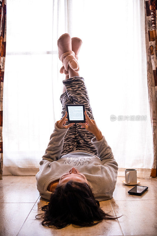 一名穿着睡衣的成年女子躺在家里客厅的地板上，一边在电子书设备上看书，一边喝着咖啡。
