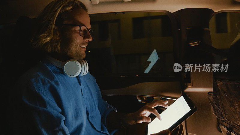 微笑的年轻人晚上坐在汽车后座上使用数码平板电脑