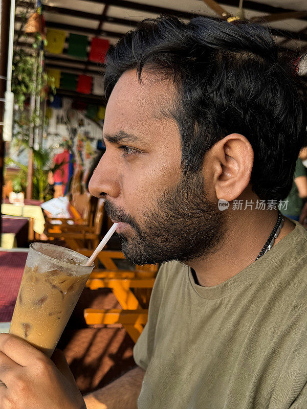 印度男子坐在露天餐厅，拿着一杯加冰块的冰咖啡饮料，用吸管啜饮，露天餐厅的背景，重点在前景
