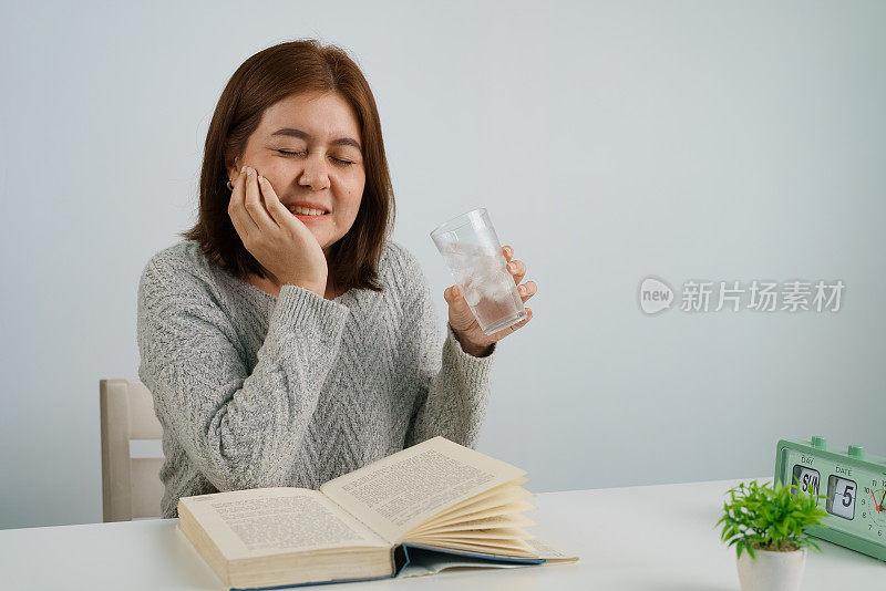 年轻的亚洲女子喝加冰的水，摸自己的脸颊，忍受牙痛，对寒冷有情绪反应。