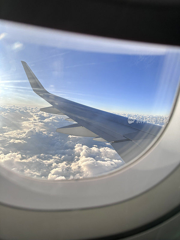 飞机机舱窗户的特写图像，部分百叶窗被拉下，透过飞机机翼的窗户看到云层上方，飞过蓬松的白云，小翼和板条，蓝天，聚焦于前景