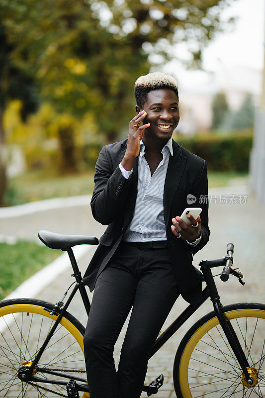 英俊年轻的非洲男子穿着西装打电话，而站在自行车工作在早上。一个商人骑着自行车去上班。