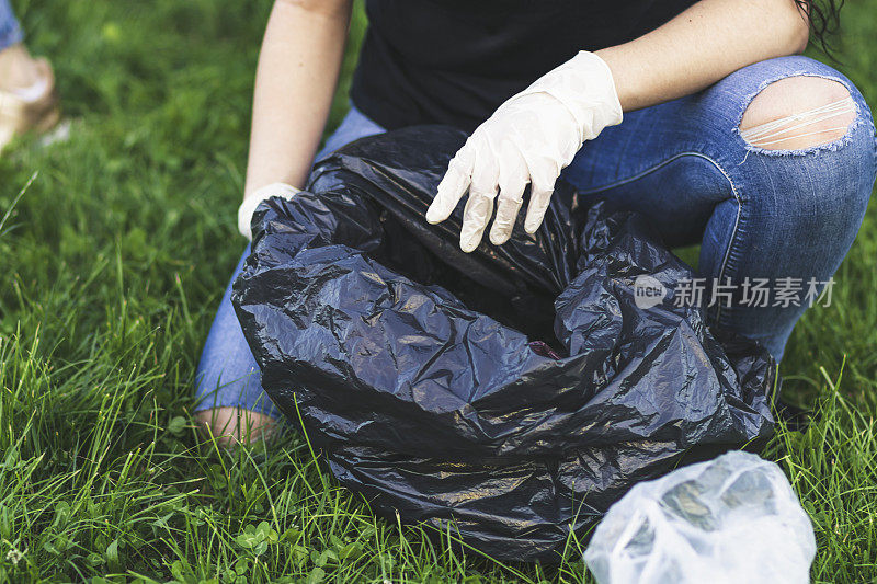 女志愿者穿着衣服在公园里捡垃圾和塑料垃圾。小女孩戴着手套，在户外把垃圾放进黑色的塑料袋里。