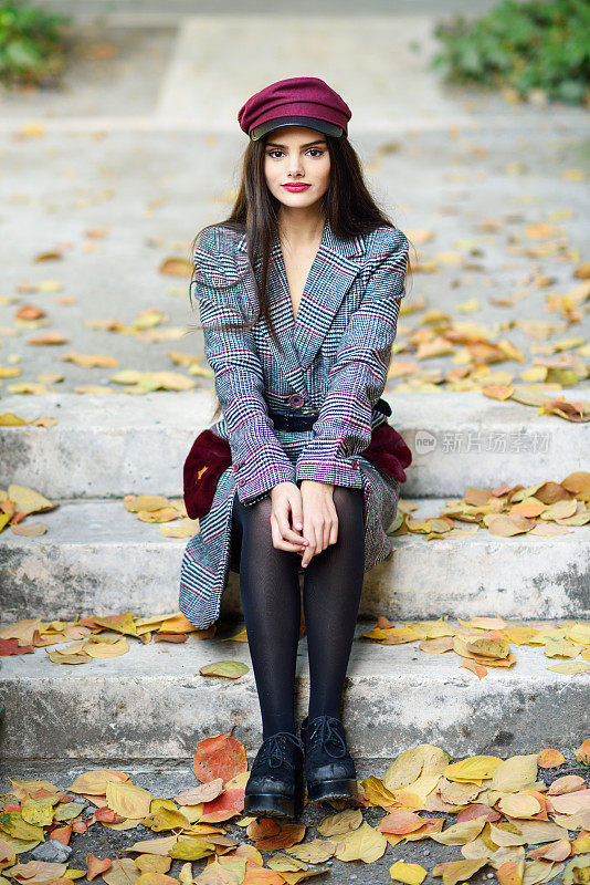年轻美丽的姑娘穿着冬衣，戴着帽子，坐在满是秋叶的台阶上