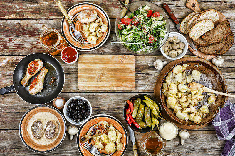 餐桌上有各种菜肴，烤鸡腿、油炸土豆配蔬菜放在煎锅里，沙拉和小吃放在木桌上，俯视图