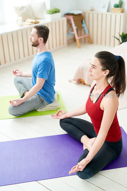 严肃冷静宁静的年轻夫妇坐在莲花体位上的运动垫和手拉手在mudra，而集中在冥想在家里