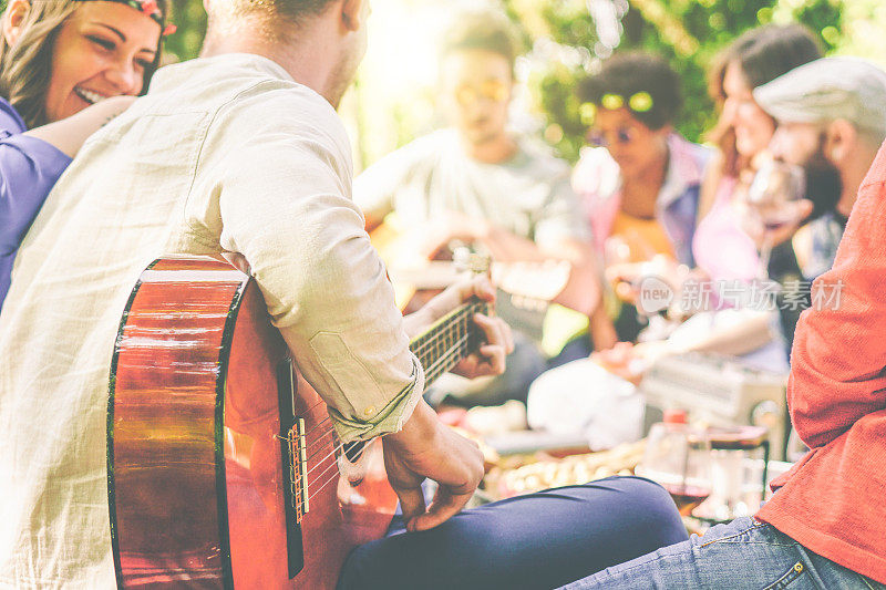 一群朋友在公园户外野餐-快乐的年轻伙伴享受野餐弹吉他，唱歌和喝葡萄酒吃食物-娱乐概念-专注于男人的手臂与吉他