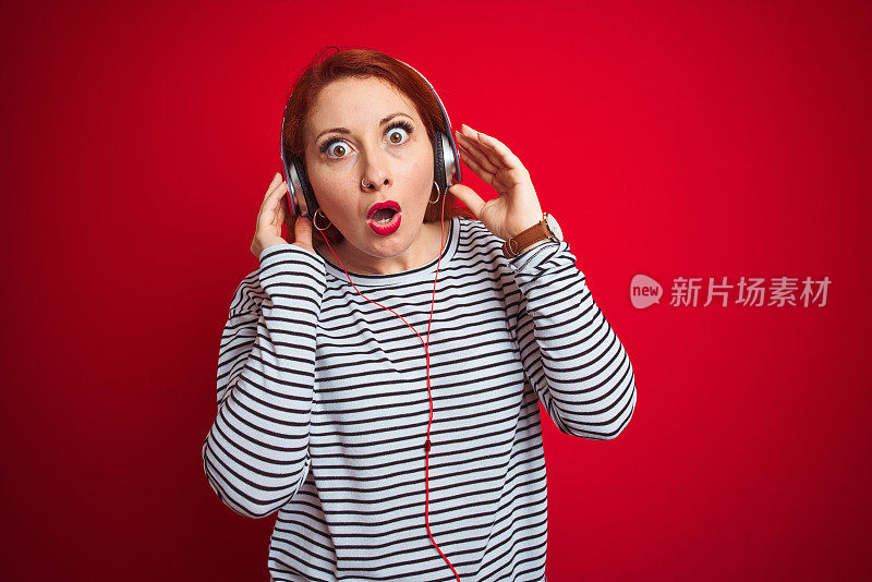 年轻的红发女子听音乐使用耳机在红色孤立的背景吓了一跳，吃惊的脸，害怕和兴奋的恐惧的表情