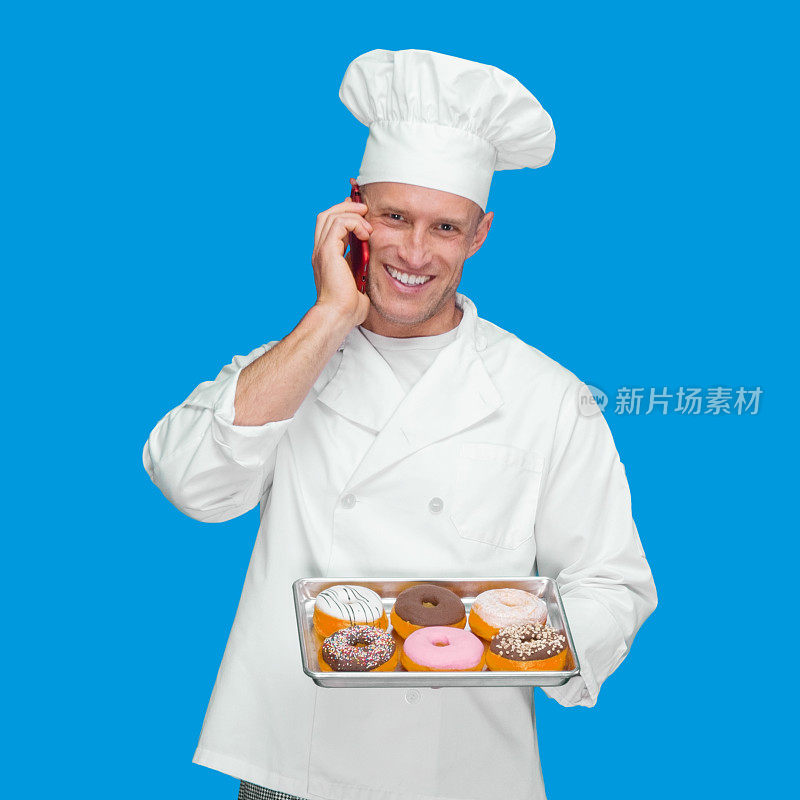 蓝色背景的白人男厨师穿着制服，拿着甜甜圈，使用手机