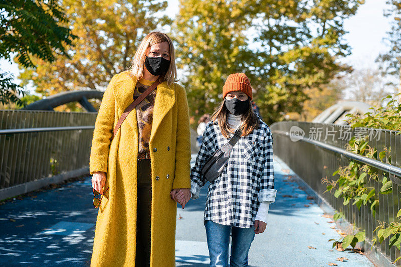 一名成年妇女和一个戴着防护面罩的女孩走在一座城市的桥上