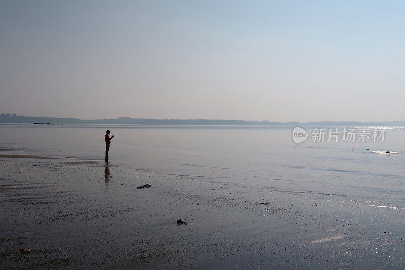一个无法辨认的人站在潮湿的沙滩上，在海浪中划桨度假，赤脚在水里走，波光粼粼的海上日落在海滩度假，帕勒姆海滩，印度果阿