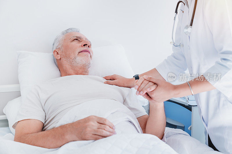 亚洲女医生手牵手，支持悲伤的老男人躺在医院的床上