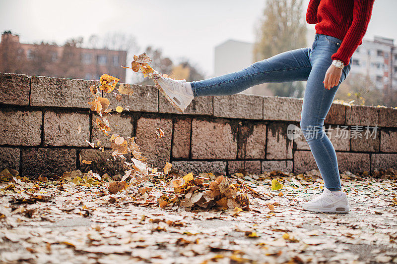 穿着白色运动鞋和牛仔裤的女人在街上踢树叶