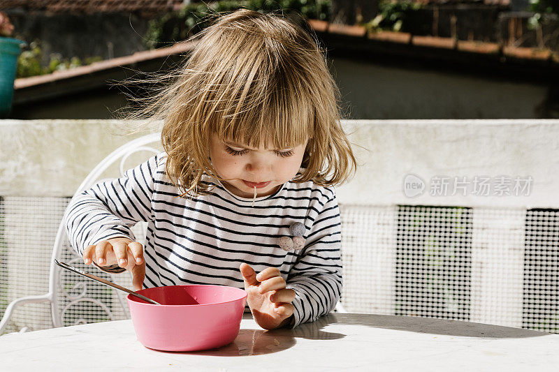 可爱的小孩坐在餐桌旁，在阳台上用粉红色的碗吃早餐，微笑着闭上眼睛。