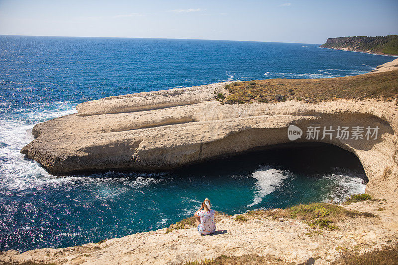 意大利撒丁岛，金发女人独自坐在天然洞穴附近的海岸上。