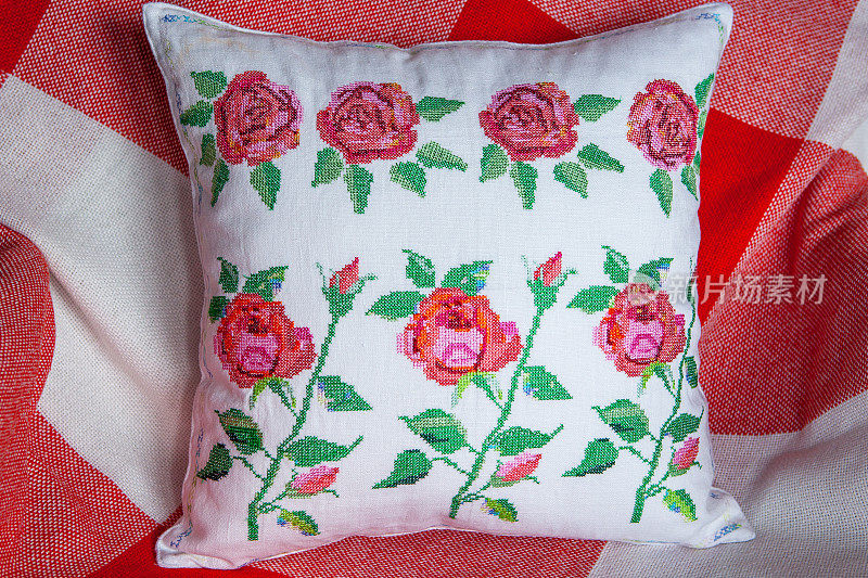 白色枕头上的玫瑰刺绣，躺在扶手椅上，铺着红白相间的毯子，背景是植物。舒适房间的内部