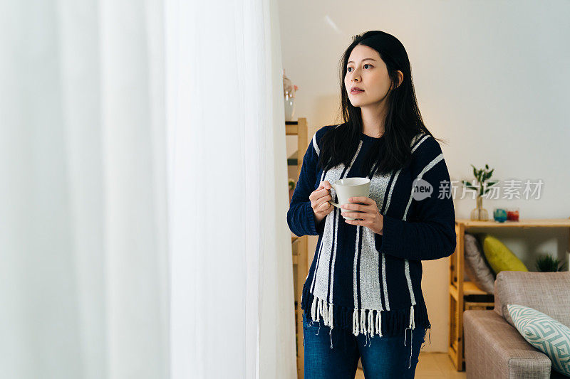 漂亮的中国女人站在窗边，拿着她的晨间咖啡。女人看着外面的天气，想着她今天的计划。